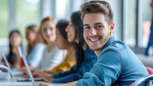 Smiling young man in a blue shirt sitting in a classroom with students working on laptops in the background.